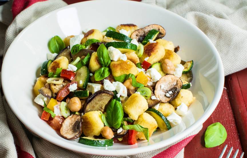 Gnocchi-Gemüse-Pfanne in einem tiefen Teller getoppt mit Feta auf einem Tisch, daneben frische Kräuter