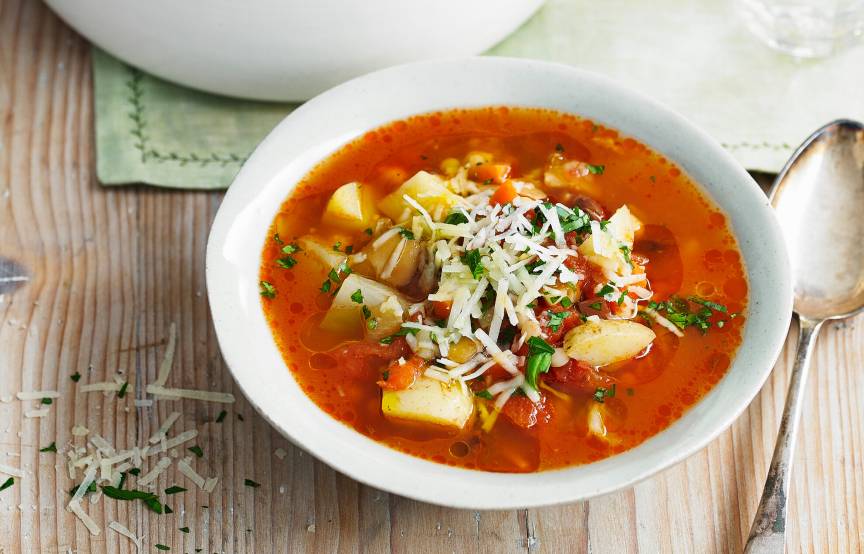 Minestrone in einer Suppenschüssel getoppt von Patmesanspänen, daneben ein voller Suppentop mit einer Kelle darin