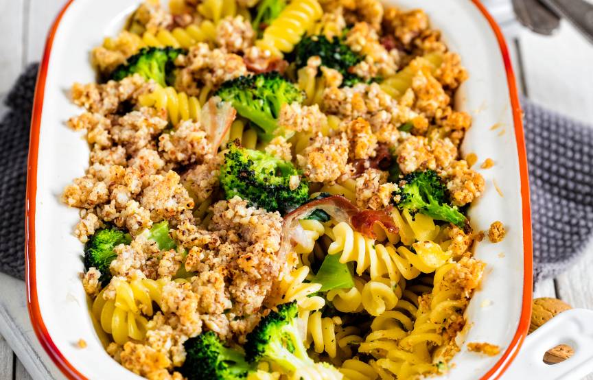 Nudelgratin mit Brokkoli, Pancetta und Mandelhaube in einer Auflaufform auf einem weißen Holztisch
