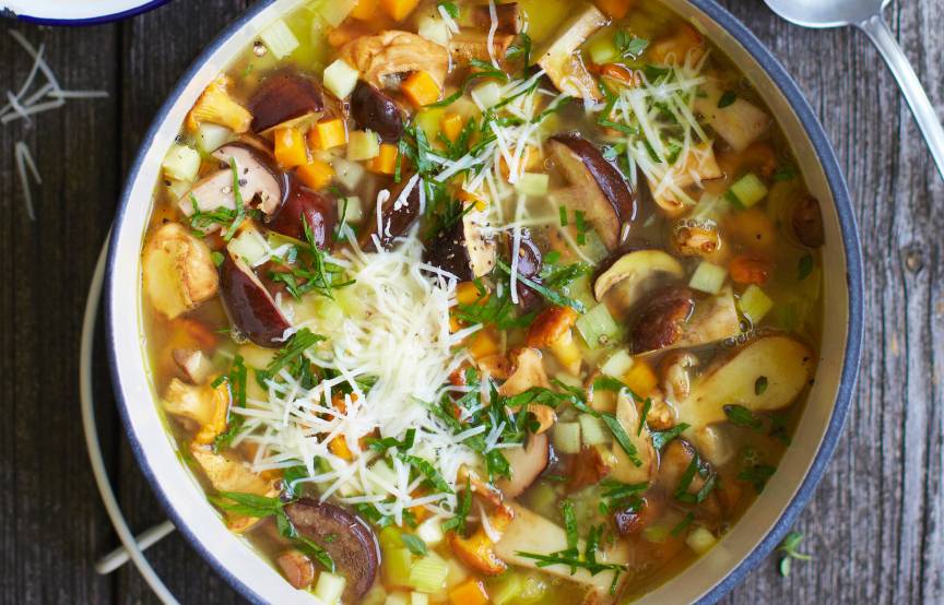 Klare Pilzsuppe mit Gemüse in einem Stieltopf auf einem Tisch, daneben gehobelter Käse und Baguette