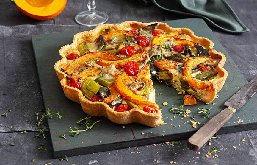 Kürbisquiche angeschnitten auf einer Schieferplatte, dahinter ein Stück Hokkaido