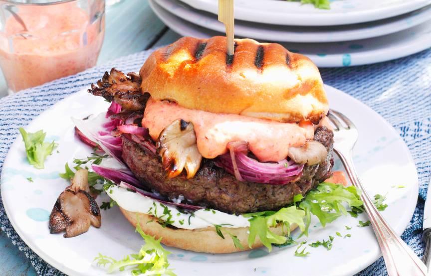 Gegrillter Burger mit Ziegenkäse und Pilzen gefüllt, zusammengehalten von einem Holzstab auf einem Teller, daneben ein Glas Mayonnaise