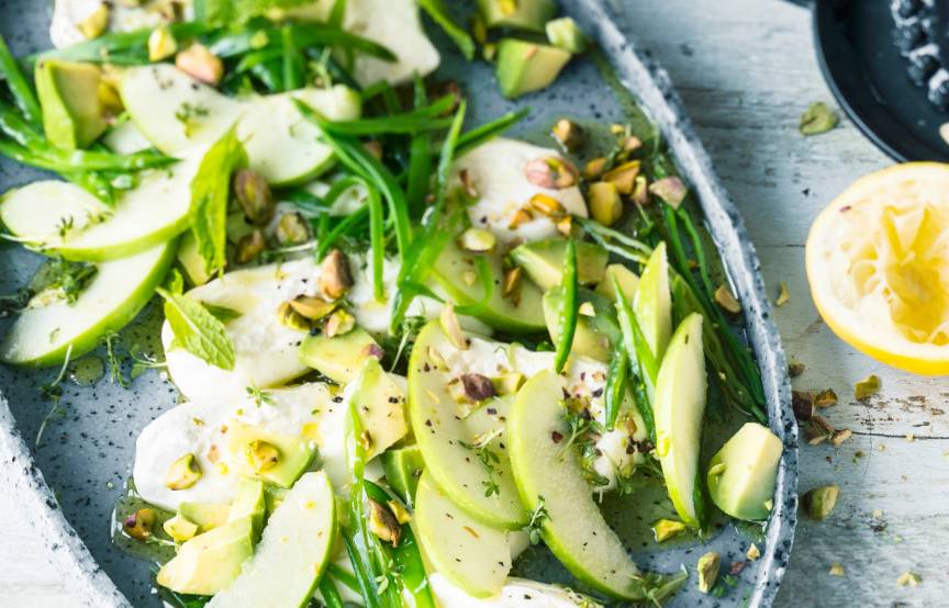 Burrata-Salat mit grünen Apfelspalten auf einem schwarzen Teller getoppt von Pistazien, daneben eine halbe Zitrone auf einem Tisch