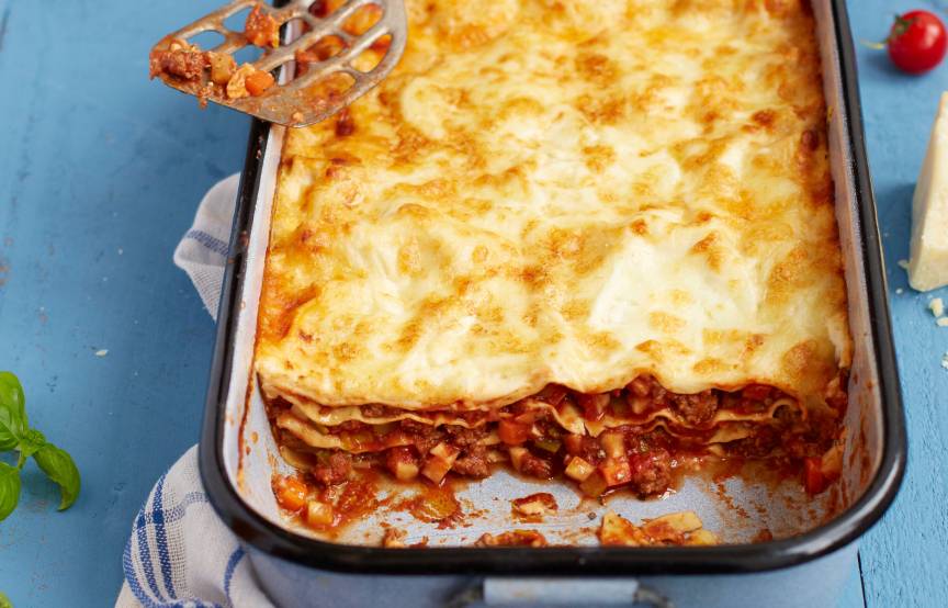 Lasagne mit Bolognese-Schichten in einer Auflaufform angeschnitten auf einem Tisch, daneben Parmesan und Tomaten