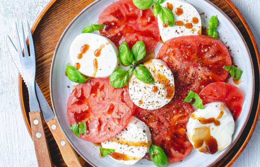 Tomaten- und Mozzarellascheiben auf einem Holzteller angerichtet als Caprese-Salat getoppt mit Balsamicodressing