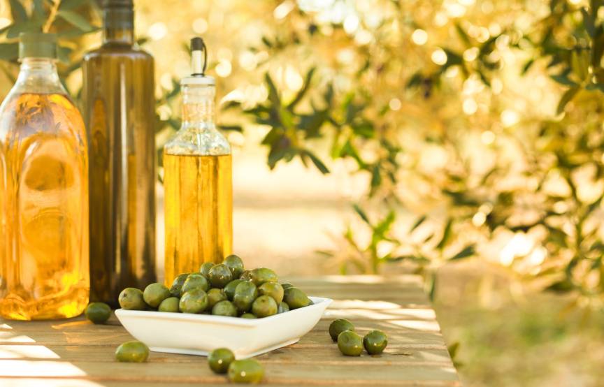 Mehrere Flaschen Olivenöl sowie grüne Oliven auf einem Holztisch vor einem Olivenhain in Italien