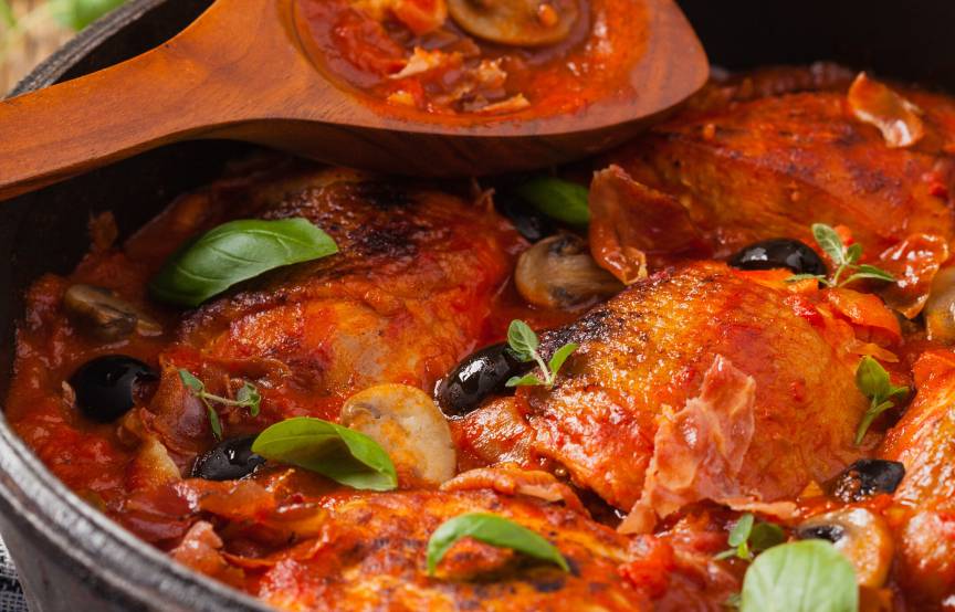 Geschmorte Hähnchenschenkel alla cacciatora in Tomatensauce in einer Pfanne garniert mit schwarzen Oliven und frischen Kräutern