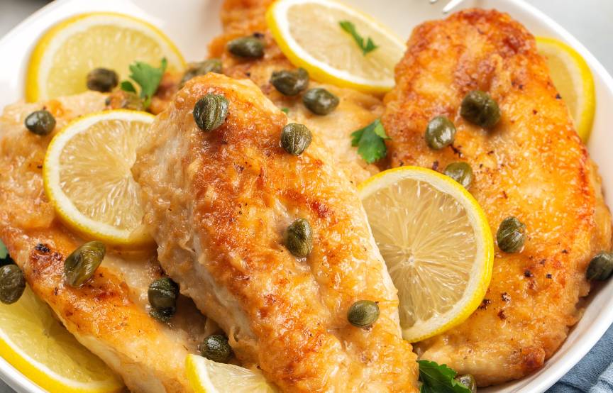 Putenbruststücke als Piccata alla milanese auf einem Teller übergossen mit Zitronen-Kapern-Sauce dekoriert mit Zitronenscheiben und frischer Petersilie