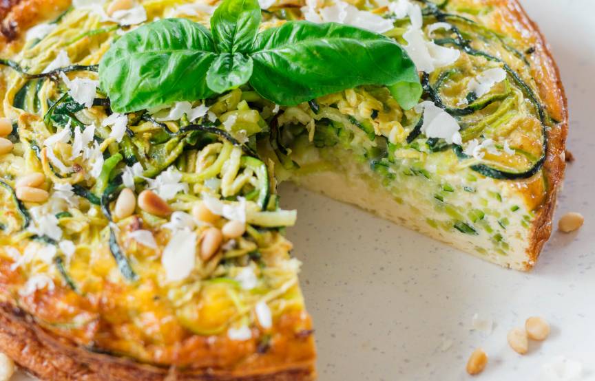 Runder Zucchini-Nudel-Kuchen angeschnitten auf einem Teller garniert mit Basilikum, daneben ein Stück Parmesan