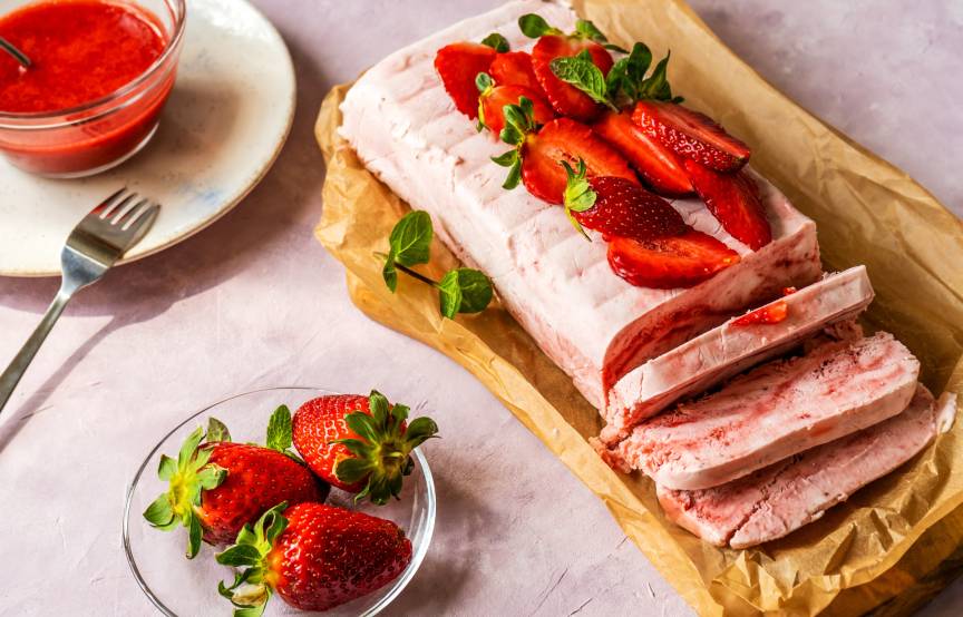 Selbst gemachtes Semifreddo angeschnitten auf einem Stück Backpapier getoppt von frischen Erdbeeren, daneben Erdbeersoße