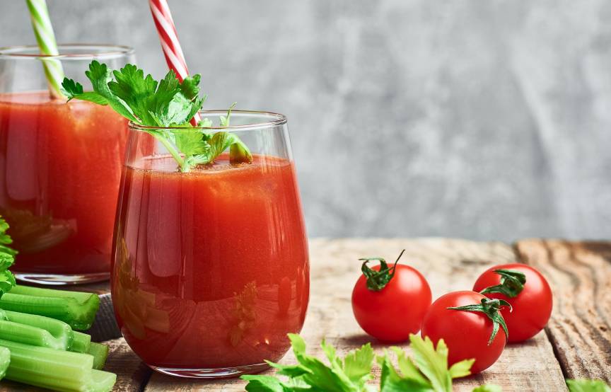 Getränk auf Tomatensaftbasis in zwei Gläsern auf einem Holztisch, daneben frische Tomaten, Sellerie und Kräuter