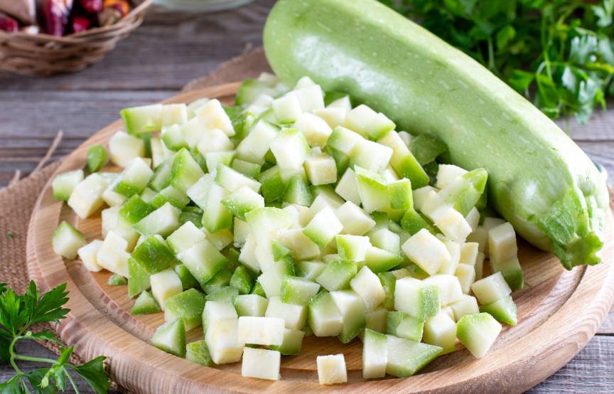 Gefrorene Zucchiniwürfel auf eine Holzbrett neben einer ganzen Zucchini