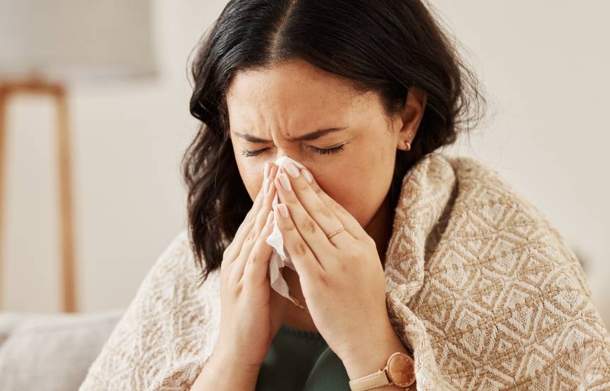 Eine Frau sitzt eingehüllt in eine Decke auf einer Couch und putzt sich die Nase
