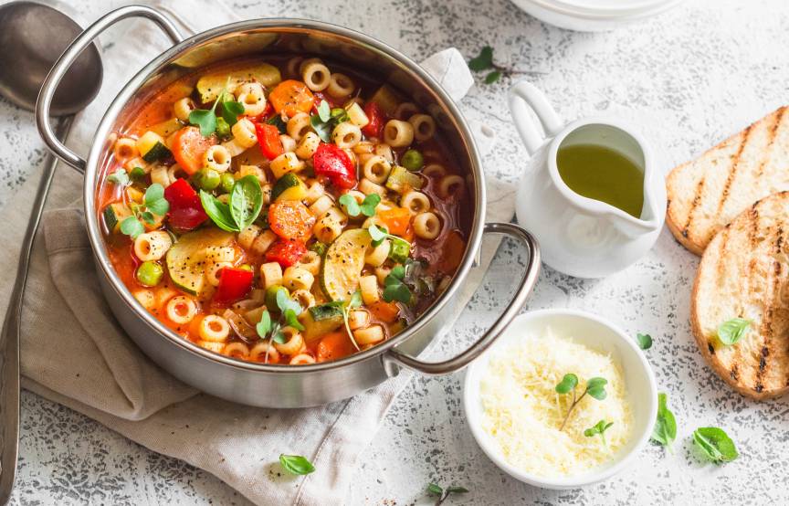 Selbstgekochte italienische Minestrone in einem Topf auf einem Tisch, daneben geröstete Ciabatta und Olivenöl