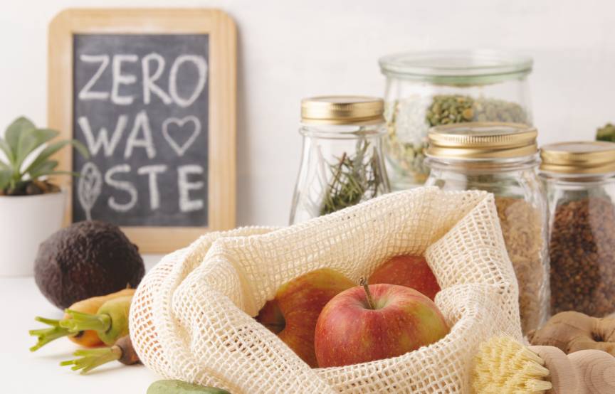 Obst und Gemüse in Stoffnetzen und Gläsern auf einem Tisch vor einer Tafel, auf der Zero Waste steht