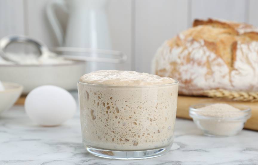 Aktiver Sauerteig in einem Weckglas auf einem Tisch mit einem Sauerteigbrot und Backzutaten