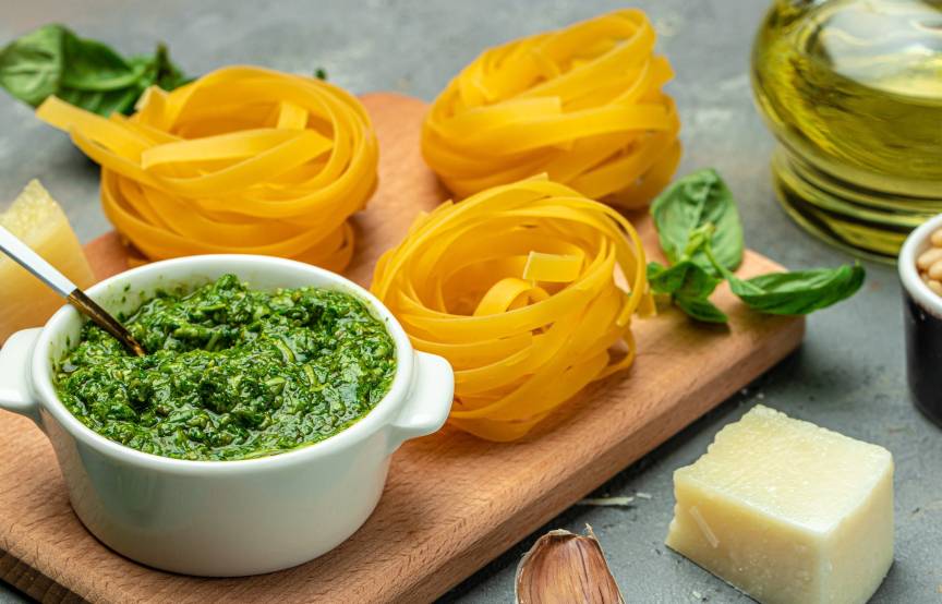 Tagliatelle auf einem Holzbrett, daneben grünes Pesto, Parmesan und andere Zutaten