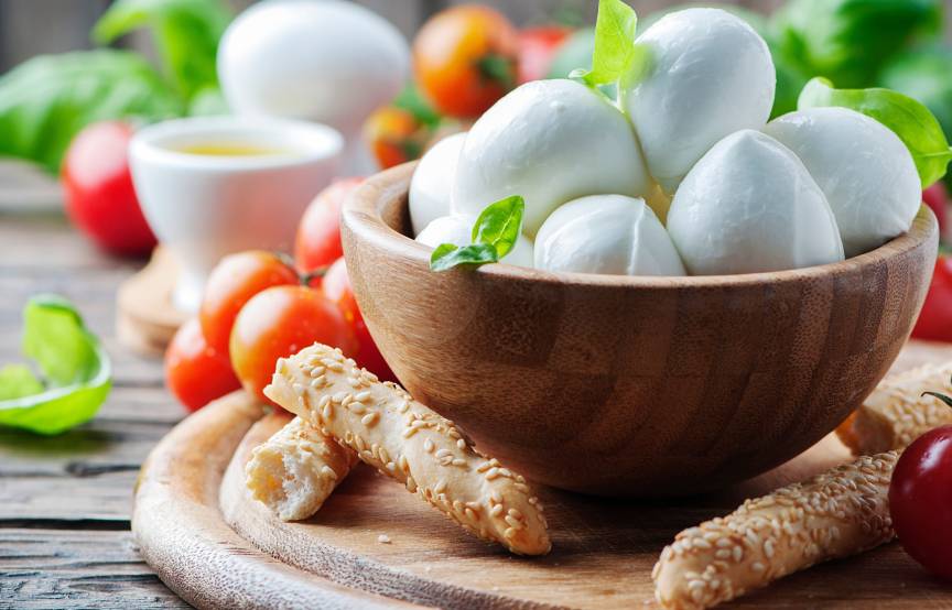 Mozzarellakugeln in einer Holzschale umgeben von Tomaten, Grissini und Basilikum