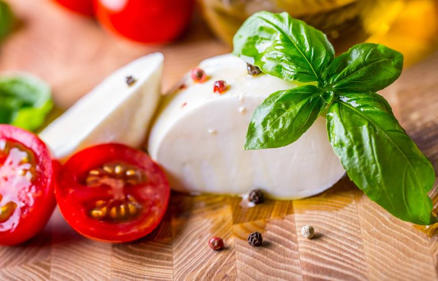 Eine angeschnittene Mozzarellakugel auf einem Holzbrett, daneben Strauchtomaten, Basilikum, Olivenöl und Pfefferkörner