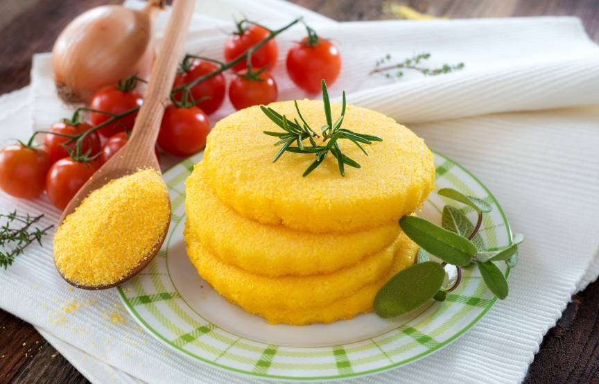Polenta in Bratlingform gestapelt auf einem Teller, daneben Tomaten, Zwiebeln und ungekochte Polenta