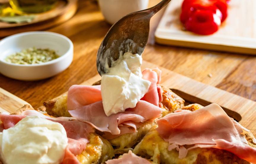 Jemand toppt eine belegte Pinsa mit Burrata, drumherum Zutaten wie Tomaten und Schinken