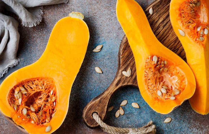 Ein aufgeschnittener Butternut Kürbis liegt ungeschält auf einem Tisch und einem Holzbrett