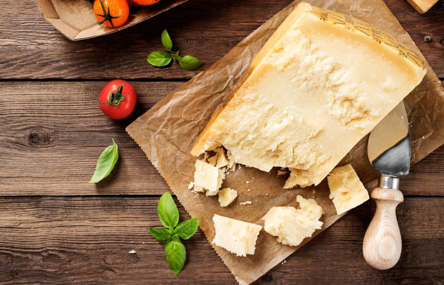 Ein Stück Parmesankäse auf einem Holzbrett, daneben Tomaten und Basilikumblätter