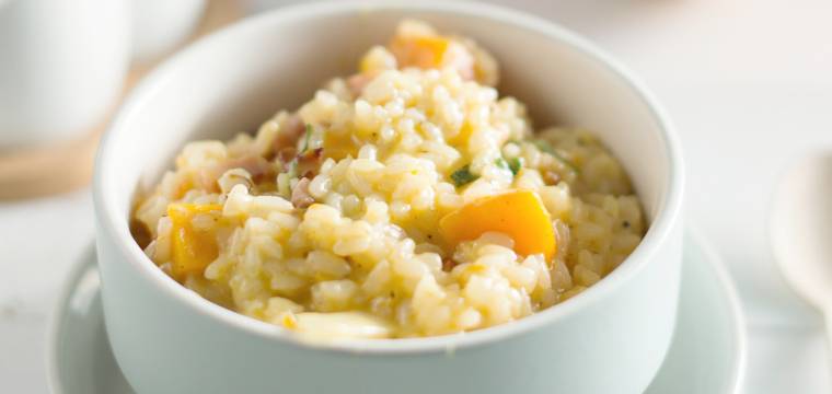 Kürbisrisotto in einer Schale auf einem Tisch, dahinter ein Stück Hokkaidokürbis