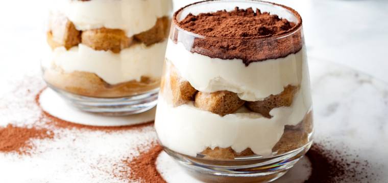 Zwei Gläser mit Tiramisu auf einer Marmorplatte bestäubt mit Kakaopulver, im Hintergrund eine Espressokanne
