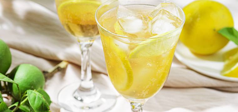 Limoncello Spritz mit Eiswürfeln in zwei Cocktailgläsern, daneben Zitronen, eine Flasche Limoncello und Zitronenmelisse auf einem Tisch