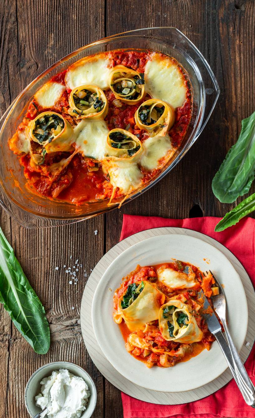 Zwei Mangold-Lasagne-Schnecken gefüllt mit Ricotta auf einem teller in Tomatensauce, daneben die Auflaufform