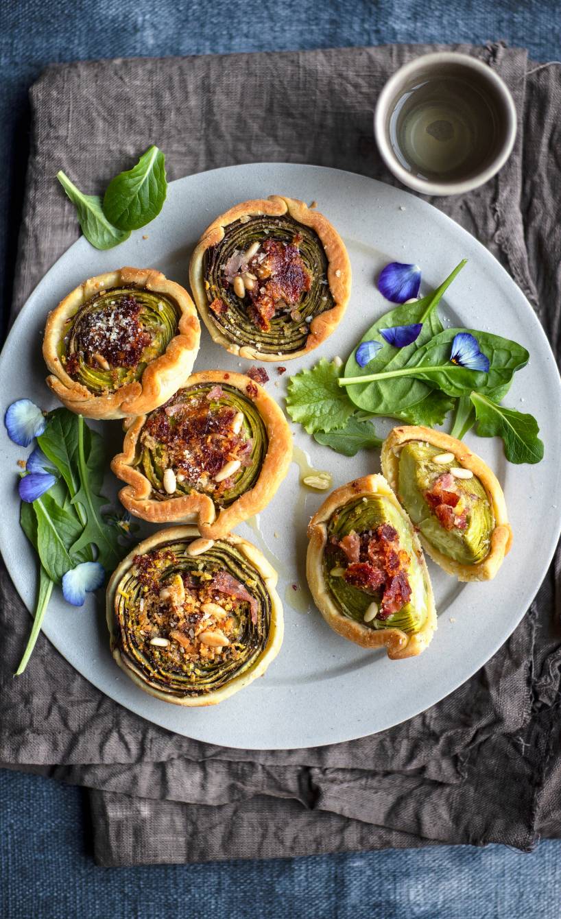 Mehrere selbstgebackene Artischocken-Törtchen aus Mürbeteig mit Parmaschinken und Käse getoppt von Pinienkernen auf einer Servierplatte