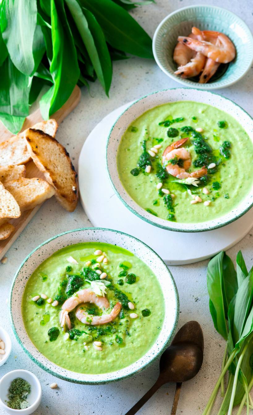 Bärlauchcremesuppe in zwei Schalen auf einem Tisch, daneben Ciabattascheiben und frischer Bärlauch