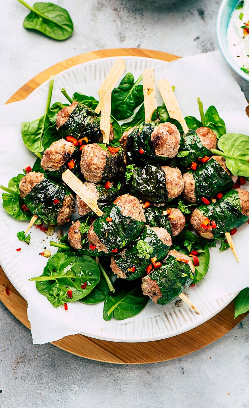 Hackfleischröllchen im Bärlauchmantel auf Spießen auf einem Salatbett, daneben Joghurtsauce auf einem Tisch