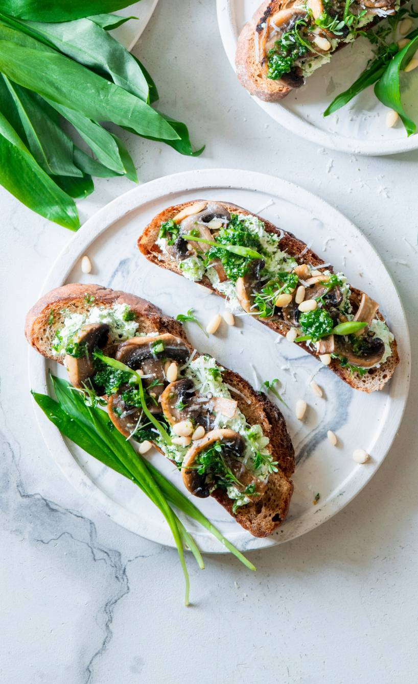 Bruschetta mit gebratenen Pilzen und Bärlauchpesto auf einem Teller, daneben frischer Bärlauch auf einem Tisch
