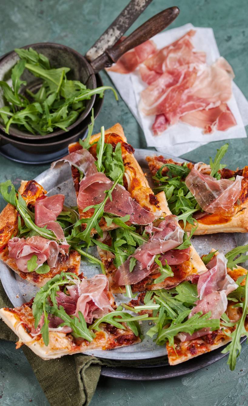 Mehrere Stücke Blätterteigpizza mit Parmaschinken, Mozzarella und Rucola angerichtet auf einem Teller