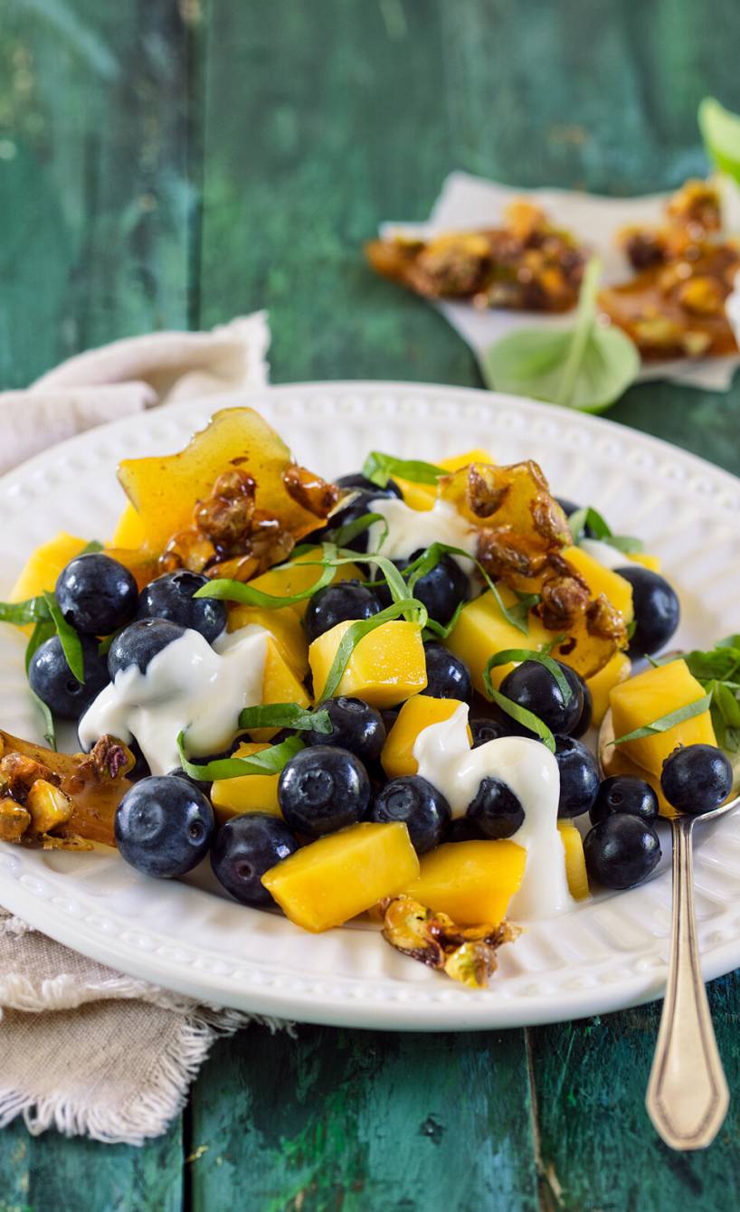 Blaubeer-Mango-Salat auf einem Dessertteller getoppt von Joghurt-Mascarponecreme und Pistazien-Krokant