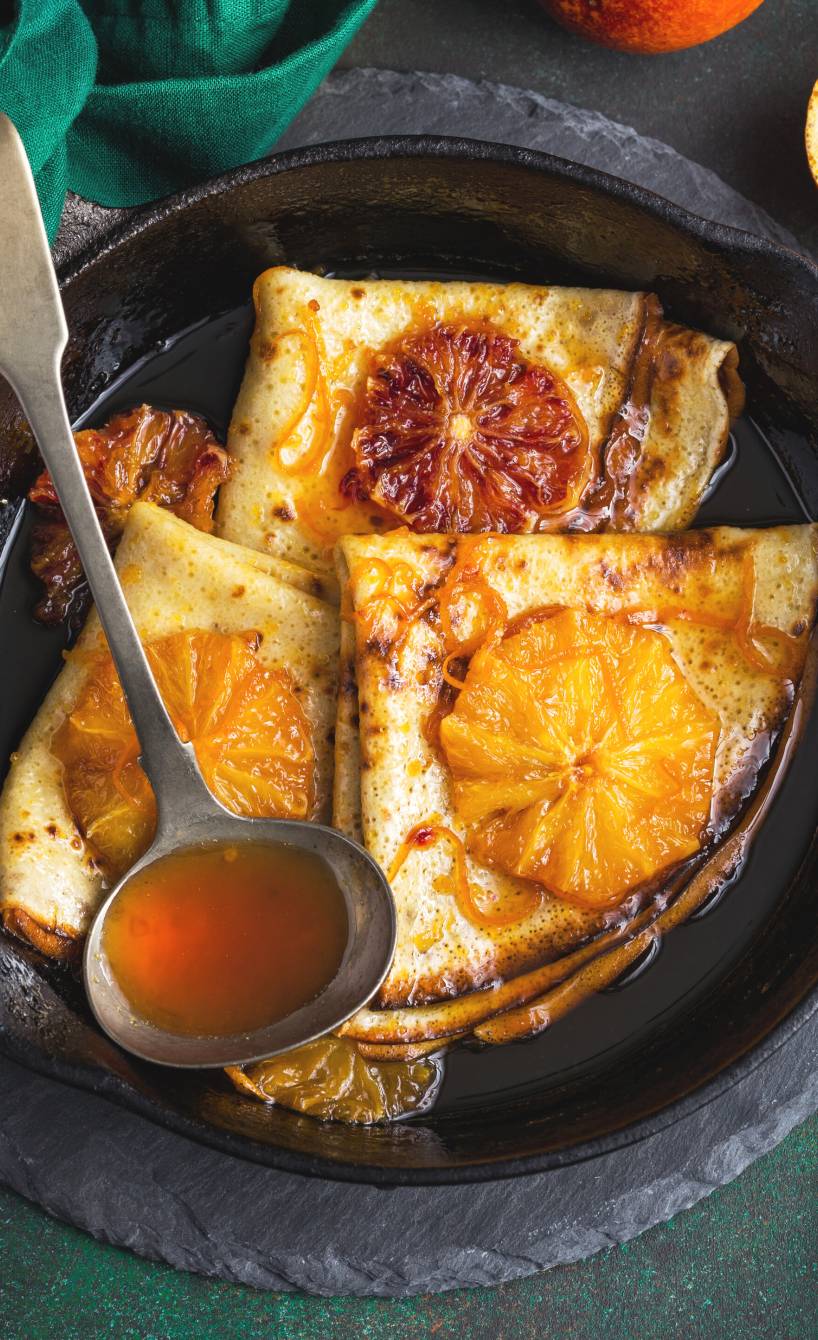 Mehrere gefaltene Crespelle auf einem Dessertteller getoppt von mit Blutorangenscheiben und Amaretto-Sauce