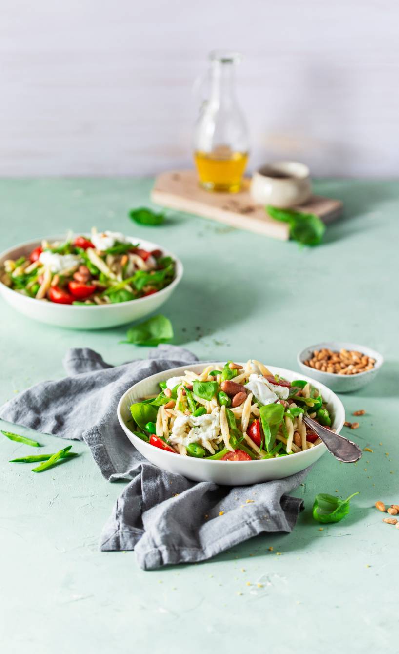 Pastasalat mit Bohnen und Zuckerschoten auf einem Teller getoppt von Büffelmozzarella, daneben Pinienkerne in einer Schale