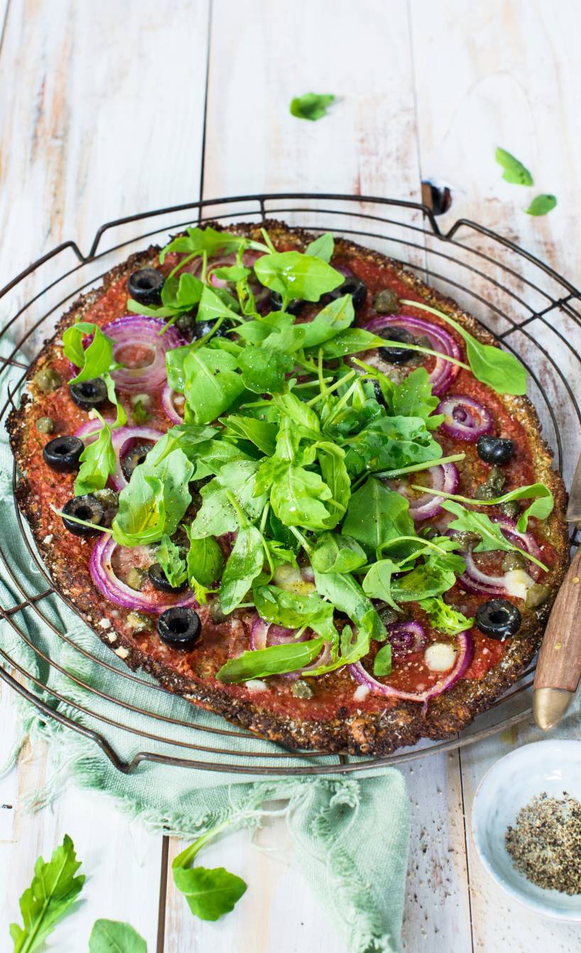 Ovale Low-Carb-Pizza mit Brokkoli-Käseboden und Belag aus Tomatensoße, Oliven, Kapern, Käse und Rucola auf einem Küchengitter
