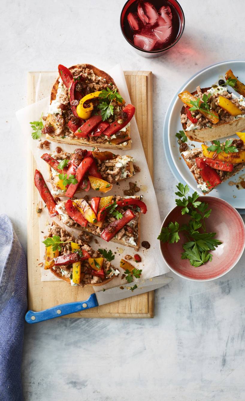 Großes Bruschetta in Streifen geschnitten auf einem Holzbrett getoppt mit Paprikasteifen, Hackfleisch und Sultaninen
