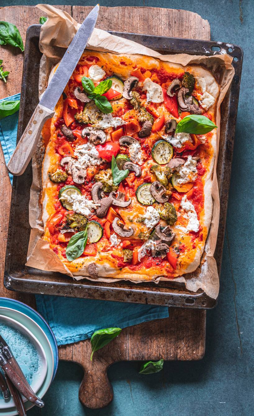 Pizza auf einem Blech belegt mit Champignons, Zucchini und Brokkoli auf einem Holzbrett