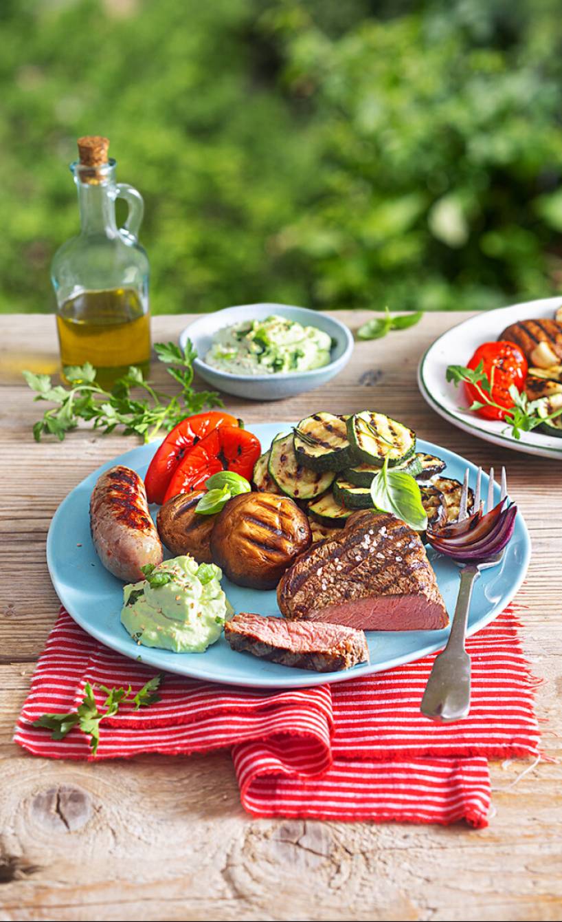 Mediterraner Grillteller mit Steak, Salsiccia, Gemüse, Avocado-Schmand-Dip und Knoblauch-Ciabatta auf einem Holztisch im Garten