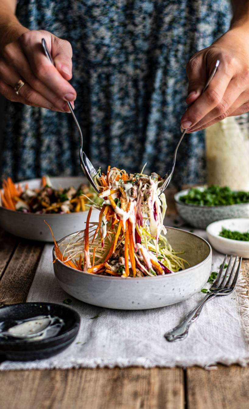 Krautsalat wird mit zwei Löffeln von einer Frau aus einer Schüssel gehoben, daneben Joghurt-Dressing in einem Glas