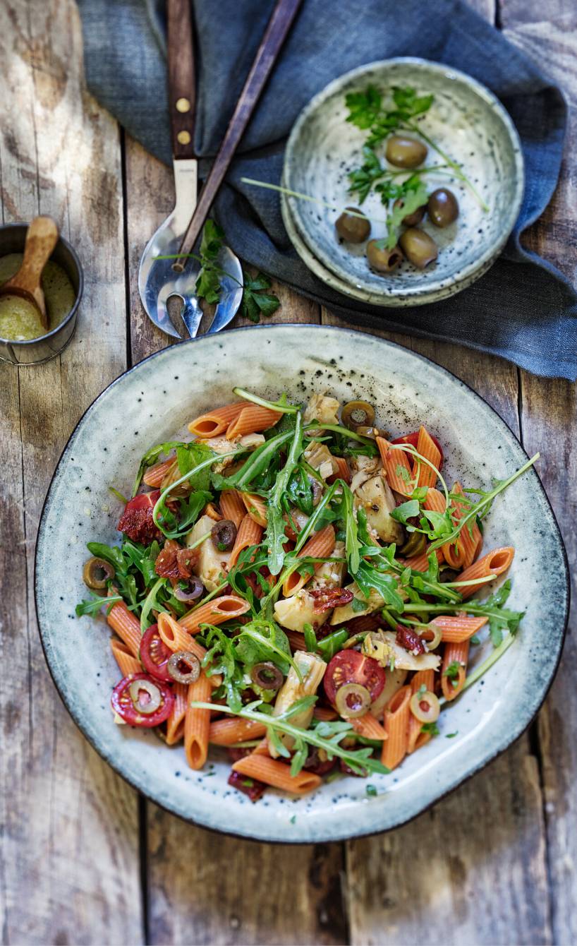 Salat aus roten Penne auf einem Teller mit Rucola und getrockneten Tomaten, daneben eine Schale mit grünen Oliven