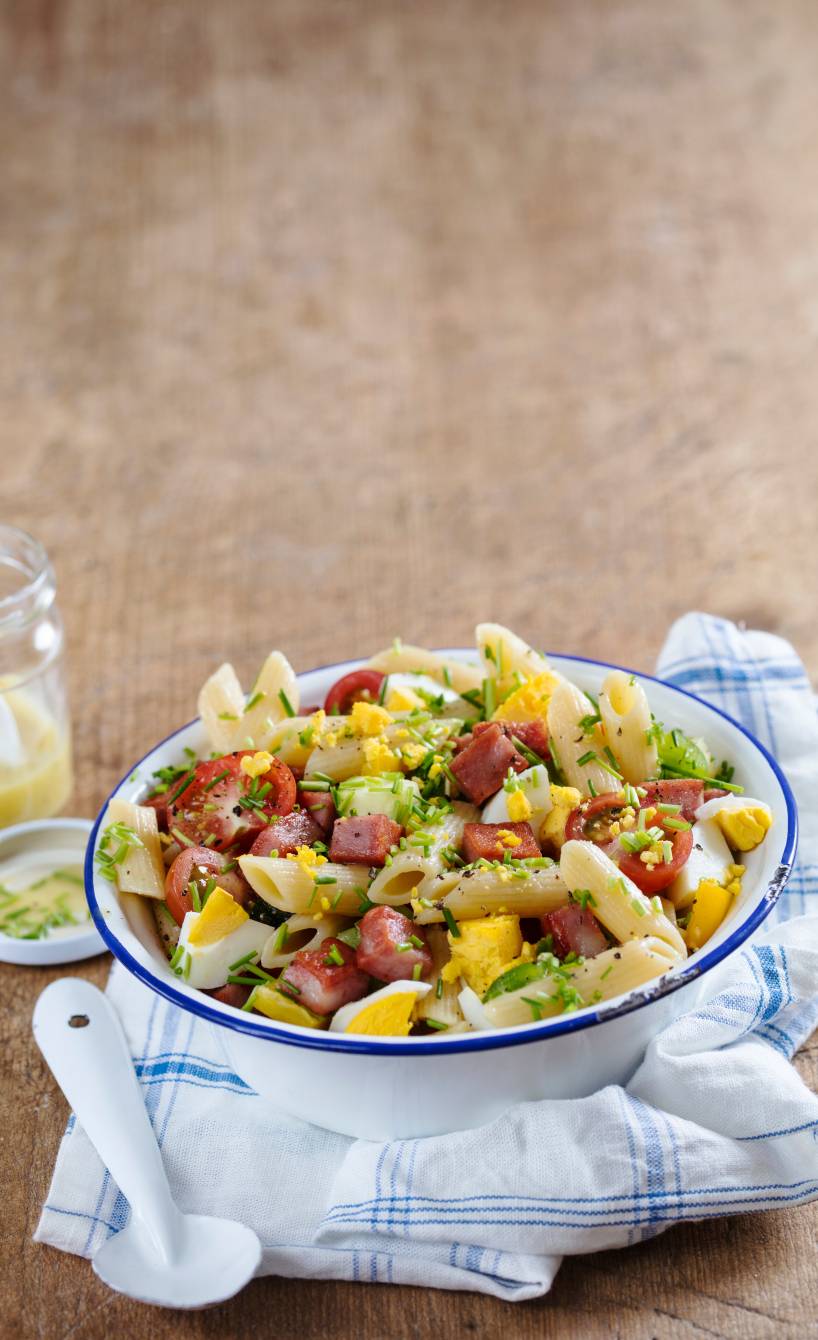 Penne-Salat in einer Schüssel getoppt mit Mortadella, Zucchini und Eierspalten, daneben Dressing in einem Glas