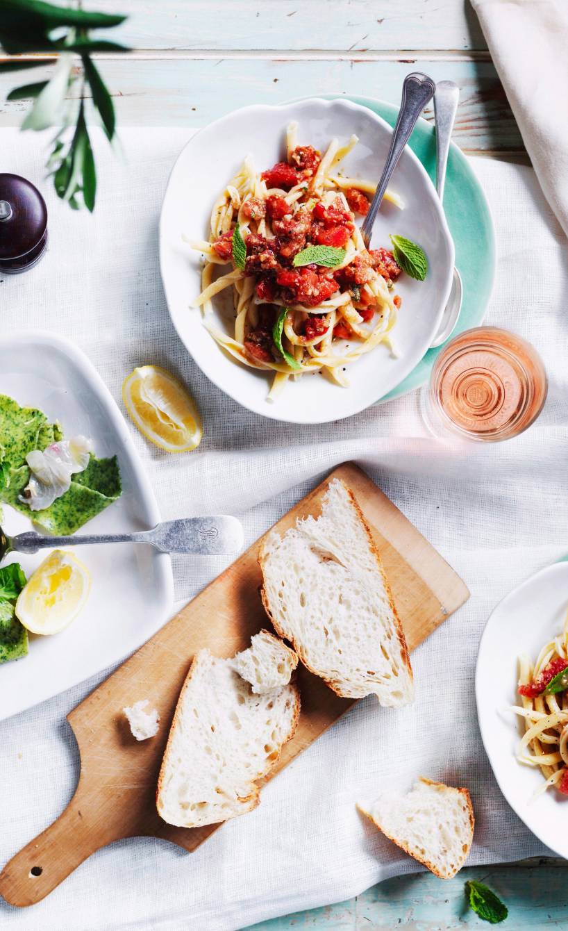 Busiate-Nudeln angerichtet auf zwei Tellern getoppt mit grünem Pesto Trapanese auf einem Tisch mit Salat und Brot