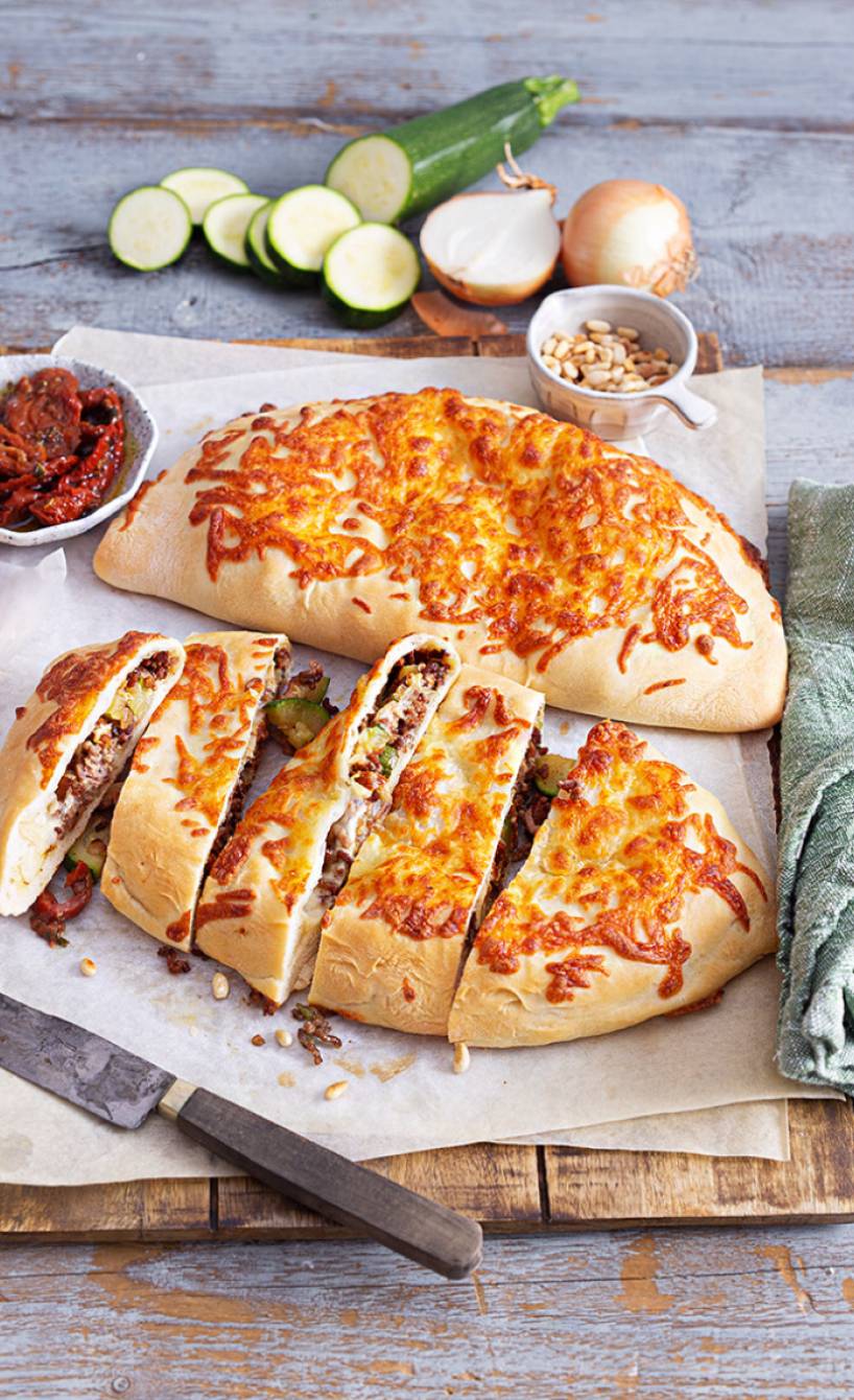 Zwei gebackene Calzones gefüllt mit Zucchini und Lammhackfleisch auf einem Holzbrett, davon eine in Streifen geschnitten