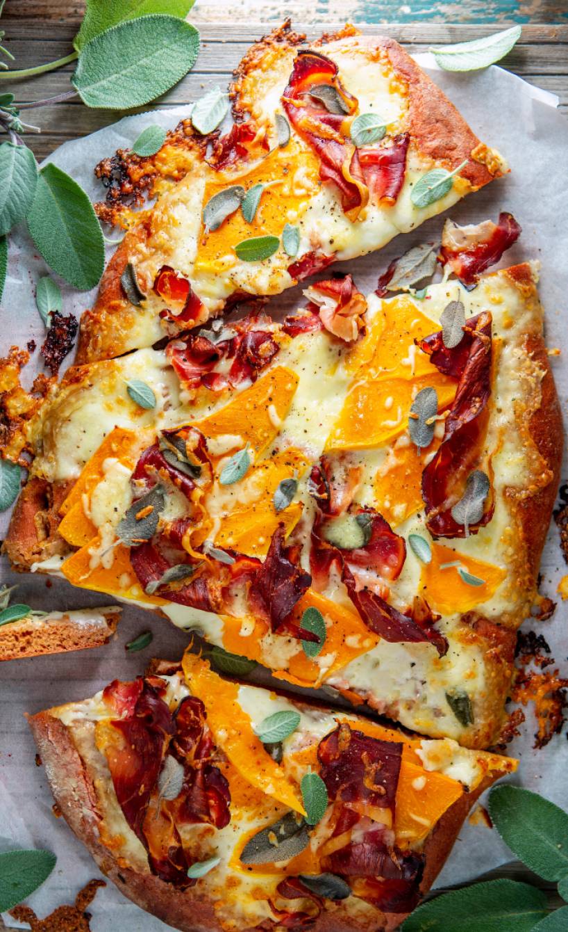 Dinkelpizza mit Butternutkürbis, Parmaschinken und Salbei in drei Teile geschnitten auf einem Tisch
