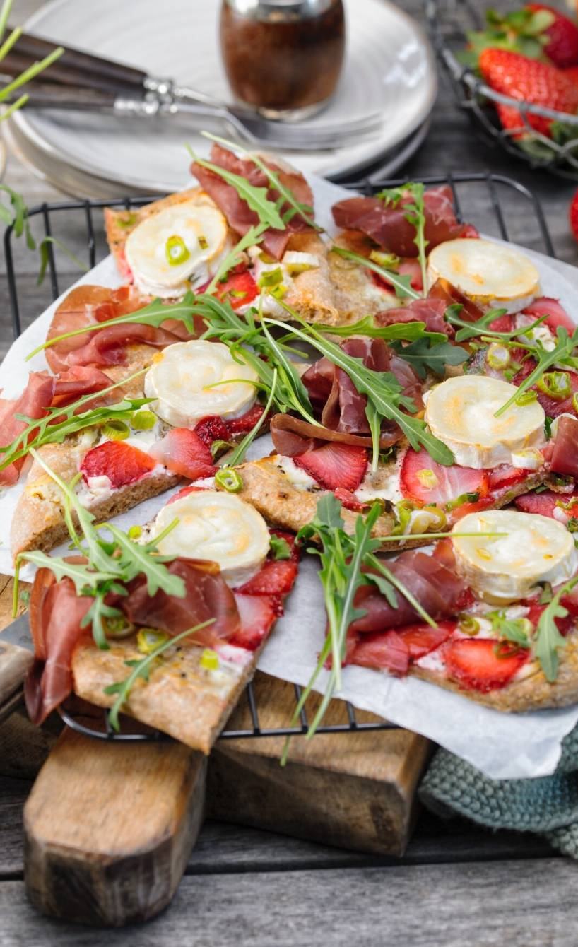 Vollkorn-Dinkelpizza belegt mit Erdbeeren, Crème fraîche, Ziegenkäse, rohem Schinken und Rucola angeschnitten auf einem Holzbrett
